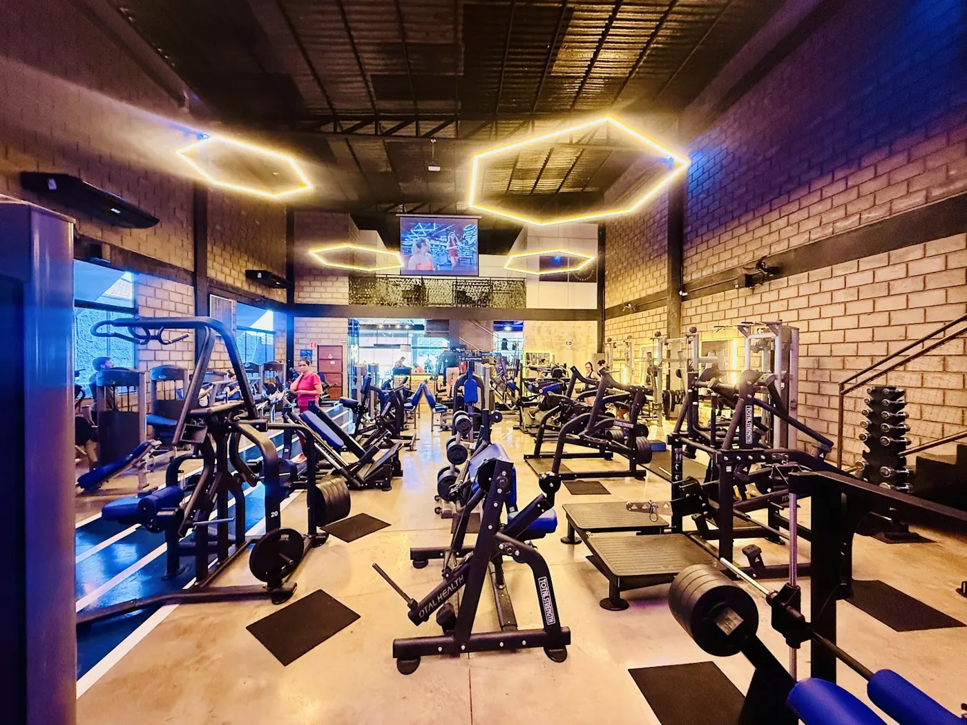 Sala de musculação da Espaço Fitness com luminárias hexagonais de LED, parede de tijolinho e aparelhos novos